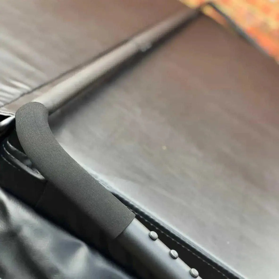 Close-up of a padded black surface and the handle of a folded HTL Cover Lifter by Hot Tub Liverpool. The handle, much like those found on hot tub accessories, is covered in a foam-like material and positioned diagonally on the left side of the image. The HTL Cover Lifter appears to be in a compact, storage position.