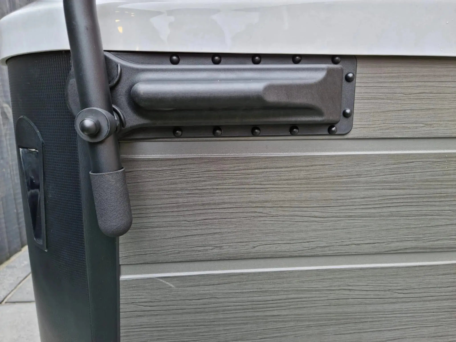 The image shows a close-up of a gray and beige hot tub exterior. There is a black handle attached to the side, resembling the HTL Cover Lifter by Hot Tub Liverpool. The texture of the hot tub's surface resembles wood grain. A black vertical panel is visible on the left side, suggesting additional hot tub accessories for convenience.