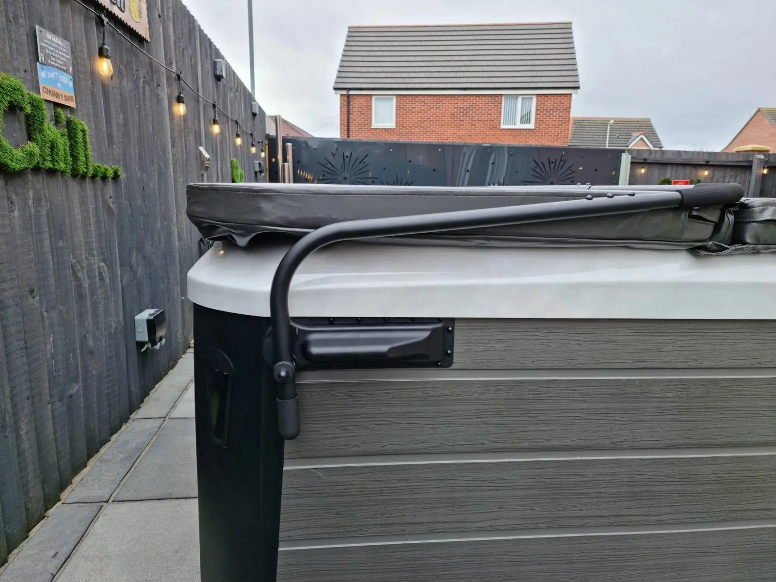 A gray and white hot tub with a Hot Tub Liverpool HTL Cover Lifter sits in an outdoor fenced area. Behind the hot tub, there is a fence adorned with string lights and decorative greenery. A brick house is visible in the background.