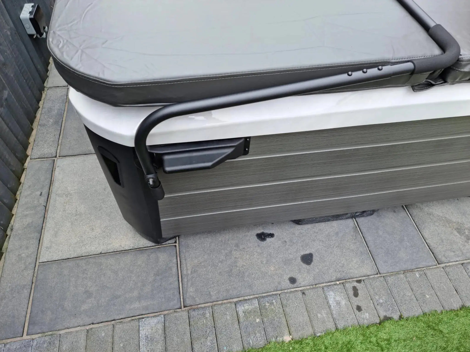 A close-up view of a hot tub showcases its partially closed gray cover. Situated on a tiled patio with artificial grass visible below, the hot tub is equipped with a Hot Tub Liverpool HTL Cover Lifter, enhancing convenience when removing the cover.