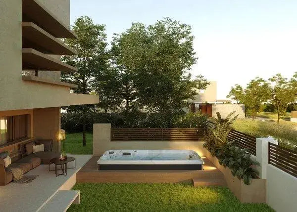 A modern backyard with a sunken Hot Tub Liverpool J-16 Powerpro™ Swim Spa, a cozy outdoor seating area with a couch and lamp, surrounded by green grass and various plants. The space is bordered by a fence, with trees and additional houses visible in the background.