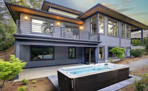 A modern two-story house with large glass windows and a spacious balcony. In front, there's a small pool or hot tub featuring the luxurious J-16 Powerpro™ Swim Spa by Hot Tub Liverpool on a patio surrounded by greenery. The sky is clearing after what appears to be a recent rain.