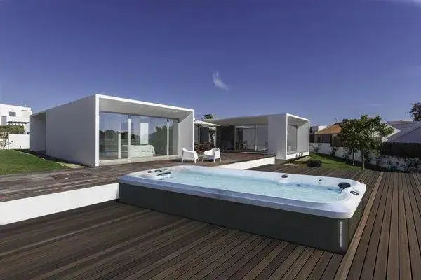 Modern outdoor space featuring a J-16 Powerpro™ Swim Spa by Hot Tub Liverpool with hydromassage seats and customizable currents, surrounded by a wooden deck. In the background, there are two contemporary minimalist white buildings with large glass windows and two white lounge chairs. The sky is clear and blue.