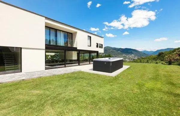 A modern, white two-story house with large windows and a spacious green lawn is shown. The property features a J-16 Powerpro™ Swim Spa on the patio with Hot Tub Liverpool jets, delivering customizable currents for ultimate relaxation. In the background, there is a scenic view of mountains under a blue sky with fluffy clouds.