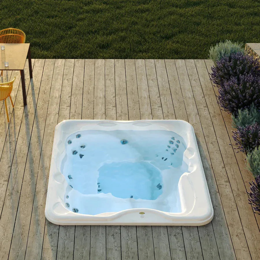 An outdoor luxurious Jacuzzi Lodge+L Holiday Let Hot Tub embedded in a wooden deck is filled with clear blue water. The deck is surrounded by lush green grass and blooming purple flowers. Nearby, a table with chairs is positioned on the deck, perfect for family holidays, and a glass of water rests on the table.