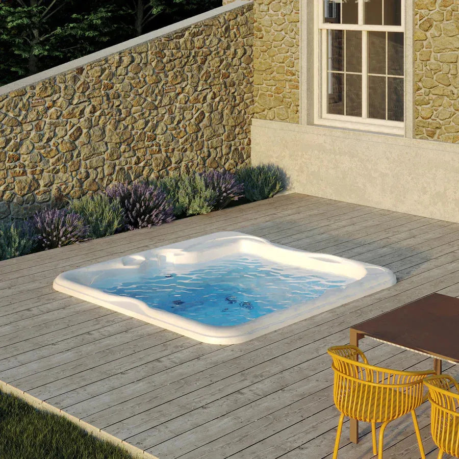 A luxurious Jacuzzi Lodge+L Holiday Let Hot Tub is set in a wooden deck next to a house with a stone exterior. Lavender plants are along the stone wall. There is a wooden table with a wicker chair nearby. The scene is outdoors, bathed in warm sunlight.