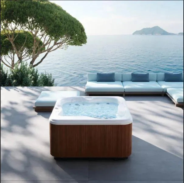 A luxurious, energy-efficient Jacuzzi Lodge+S Holiday Let Hot Tub by Jacuzzi sits on a spacious deck overlooking a calm, expansive body of water. In the background, there's comfortable seating with light blue cushions and a tree offering partial shade. An island is visible in the distance under a clear sky.