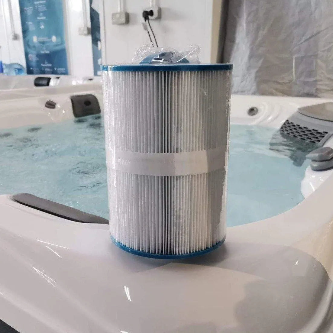 A cylindrical LHT Filter - Medium by Hot Tub Liverpool with blue end caps stands upright on the edge of a hot tub, emphasizing the importance of hot tub maintenance. The hot tub is filled with water, and a wall with a power outlet is visible in the background. The filter, wrapped in clear plastic, suggests readiness for annual filter replacement.