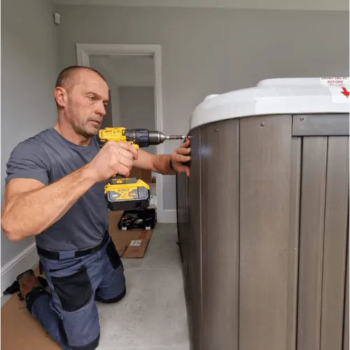 A man kneels on the floor while using Hot Tub Liverpool Repairs to repair a large gray and white piece of equipment, possibly an out of warranty tub. He is in a brightly lit, indoor space with white walls and some scattered tools on the floor nearby.