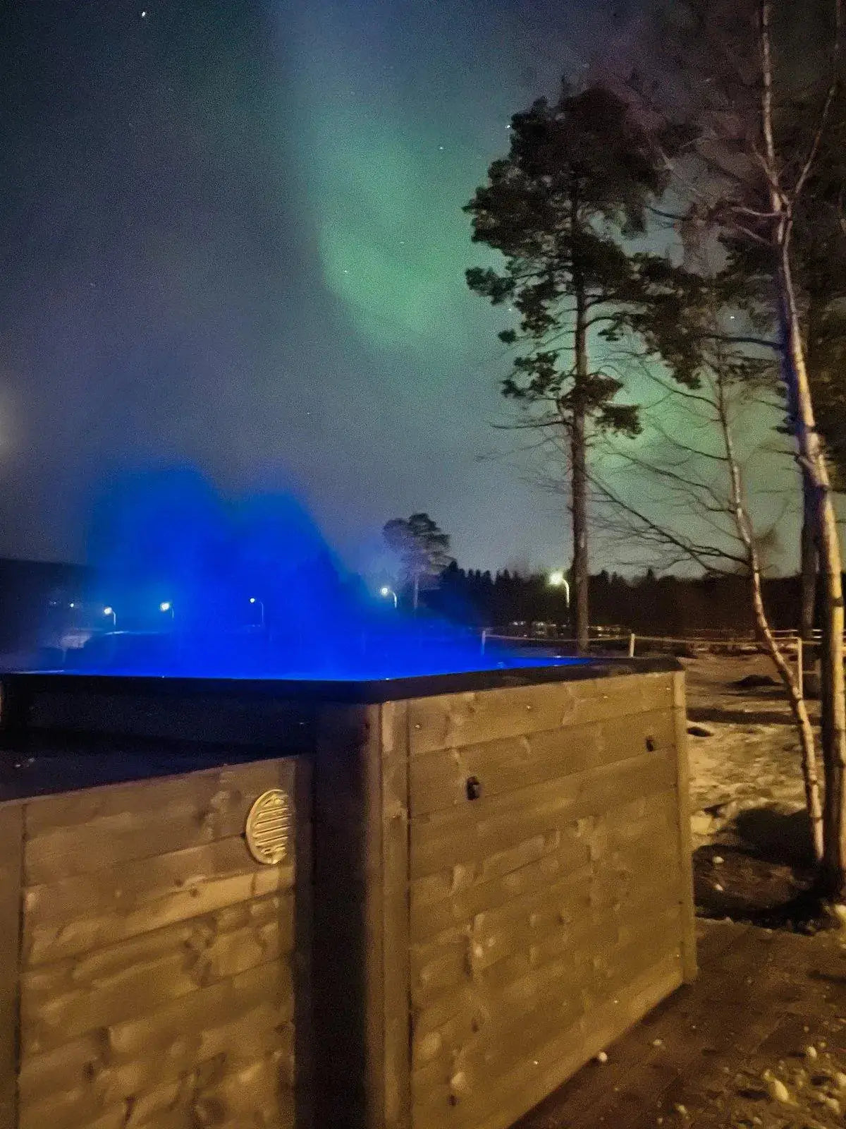 A Rexener Polar Wooden Hot Tub - Made in Finland by Hot Tub Liverpool emits steam in the foreground, illuminated by blue light. Tall trees stand nearby, with the northern lights casting green hues across the night sky. Snow covers the ground, creating a serene winter scene.