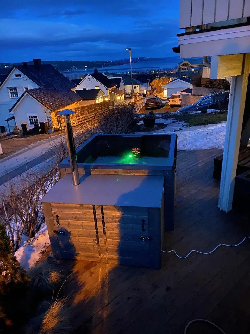 A handmade Rexener Polar Wooden Hot Tub - Made in Finland by Hot Tub Liverpool with a chimney is on a deck overlooking a snow-covered residential neighborhood at dusk. The hot tub is lit from within, casting a green glow. Houses and a body of water are visible in the background under a darkening sky.