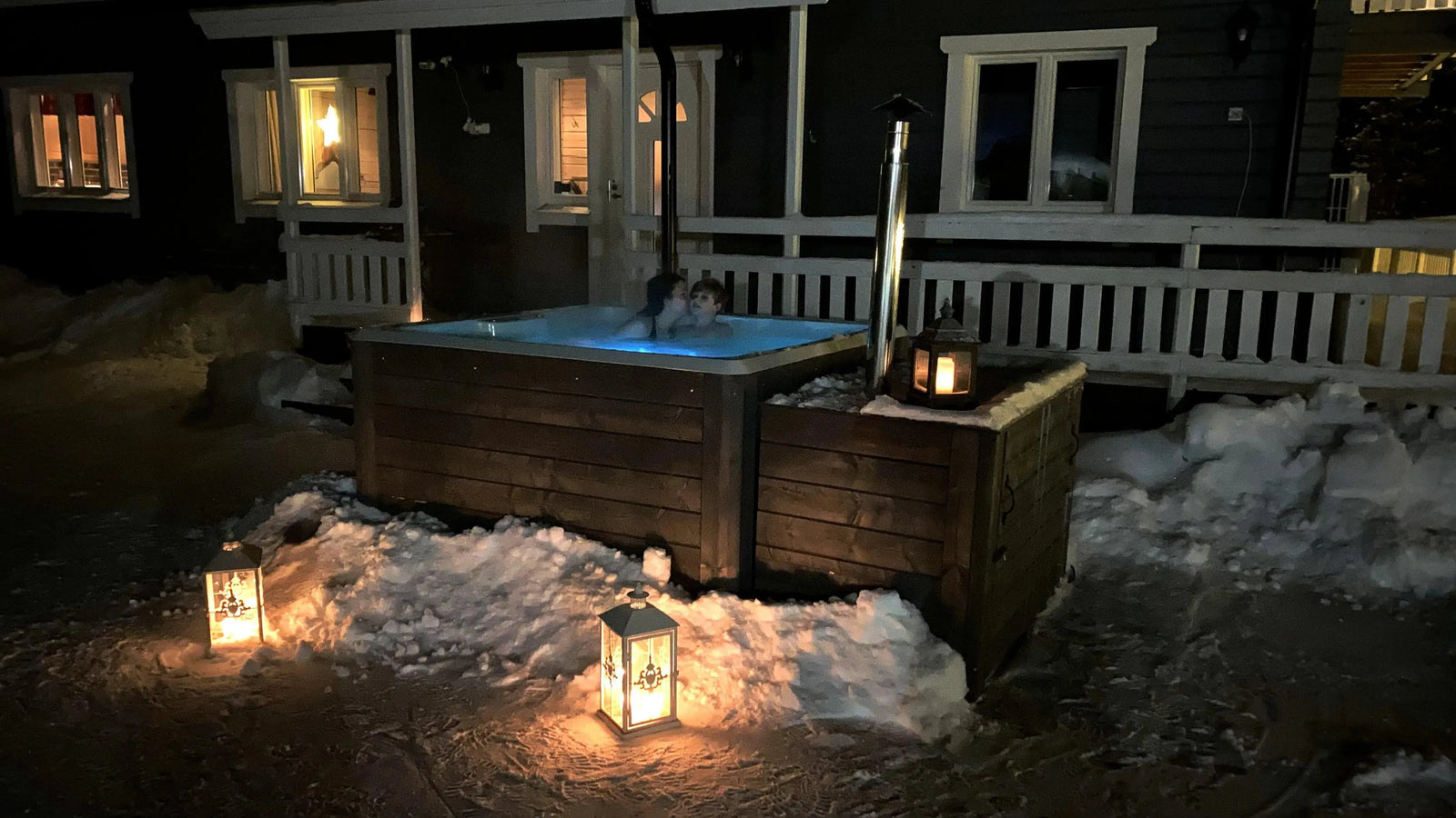 A cozy handmade Rexener Polar Wooden Hot Tub - Made in Finland sits outside a snow-covered cabin at night. Warm light from lanterns illuminates the scene. Steam rises from the hot tub, heated by a Hot Tub Liverpool water heater, where a person is relaxing. The cabin’s glowing windows add to the warm and inviting atmosphere.
