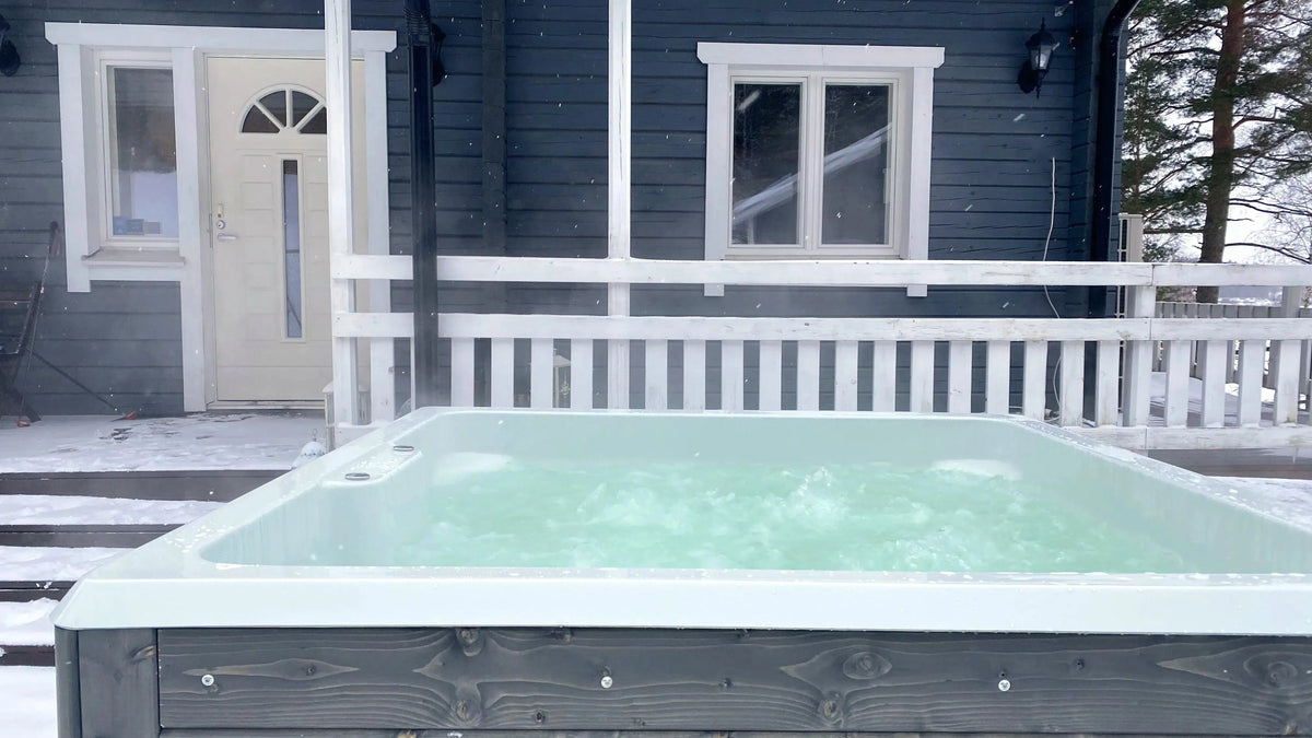 A Hot Tub Liverpool Rexener Polar Wooden Hot Tub - Made in Finland filled with water sits on a snow-covered wooden deck in front of a dark blue house with white trim. Steam rises from the hot tub, creating a cozy contrast to the wintry surroundings. The house has a window and a closed door visible.