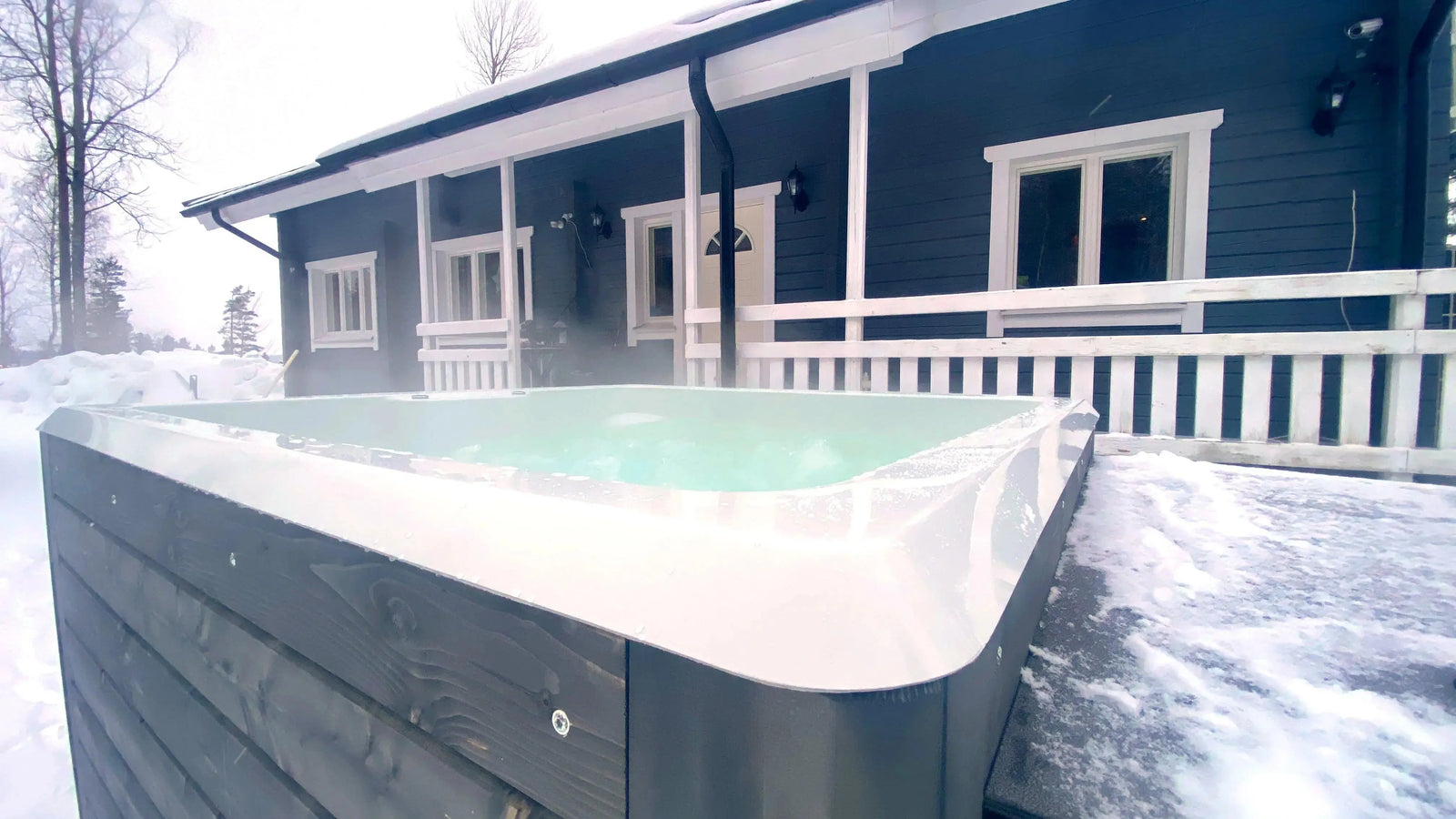 A steaming Rexener Polar Wooden Hot Tub - Made in Finland in a snowy backyard, positioned in front of a gray wooden house with white trim. The house has a covered porch with railings and windows facing the hot tub. Light snow covers the ground and the hot tub's edges, heated by an efficient Hot Tub Liverpool water heater.