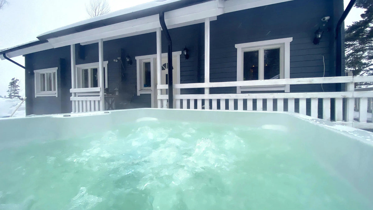 A modern cabin with a gray exterior and white trim is visible in the background, with a patio and railing. The foreground features a Rexener Polar Wooden Hot Tub - Made in Finland by Hot Tub Liverpool filled with bubbling water from the Rexener PR200 water heater, creating a relaxing and inviting atmosphere. Snow surrounds the area.