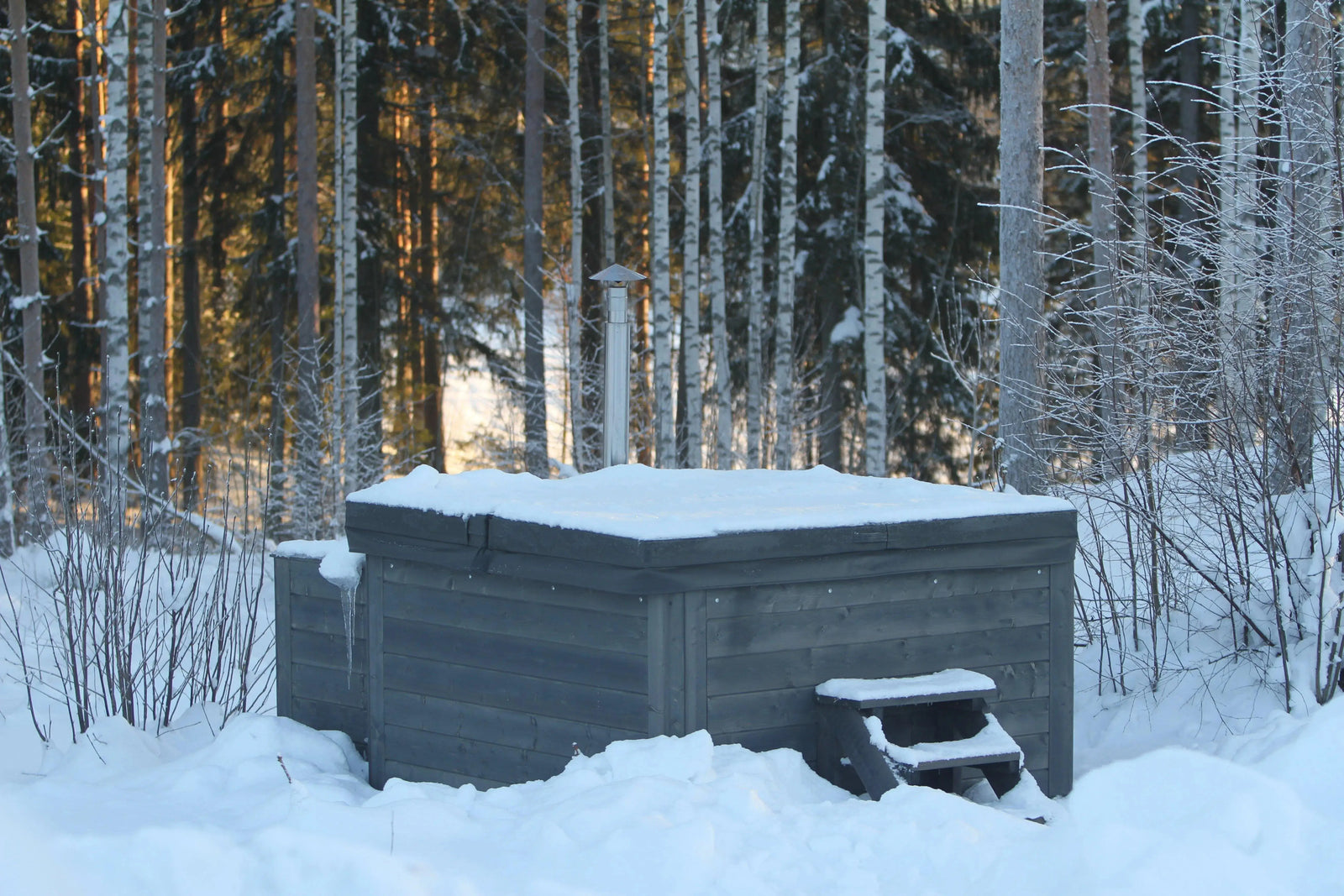A Hot Tub Liverpool Rexener Polar Wooden Hot Tub - Made in Finland covered in snow sits in a snowy forest clearing. The surrounding trees are dusted with snow, and gentle sunlight filters through the woods, casting a serene winter atmosphere. Snow-covered steps lead up to the hot tub.