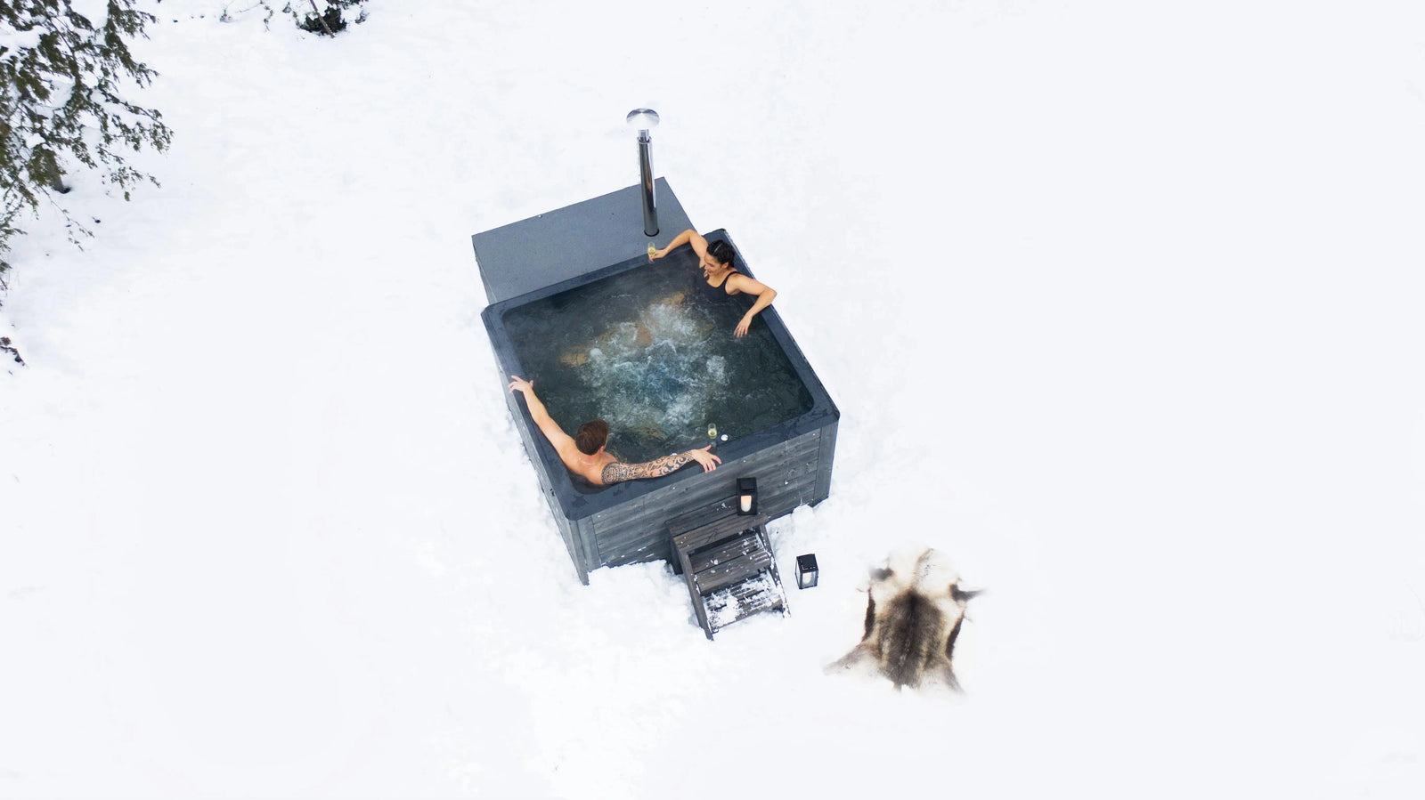 Two people are relaxing in a handmade hot tub surrounded by snow. One person with a tattoo leans on the edge, while the other stretches their arms out. A firewood storage and animal pelt are visible nearby, and tall trees frame the snowy landscape, all heated perfectly by a Hot Tub Liverpool Rexener Polar Wooden Hot Tub - Made in Finland.
