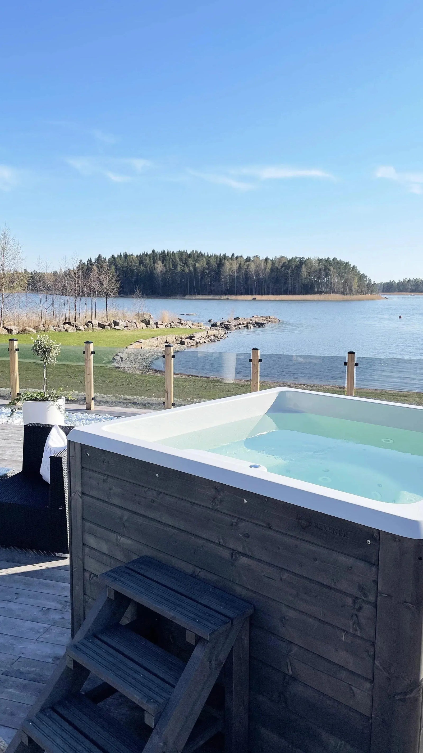 A **Hot Tub Liverpool Rexener Polar Wooden Hot Tub - Made in Finland** with clear water is situated on a wooden deck overlooking a serene lake surrounded by forested land. A wicker chair is placed nearby, and the sky is clear with bright sunlight illuminating the tranquil scene.