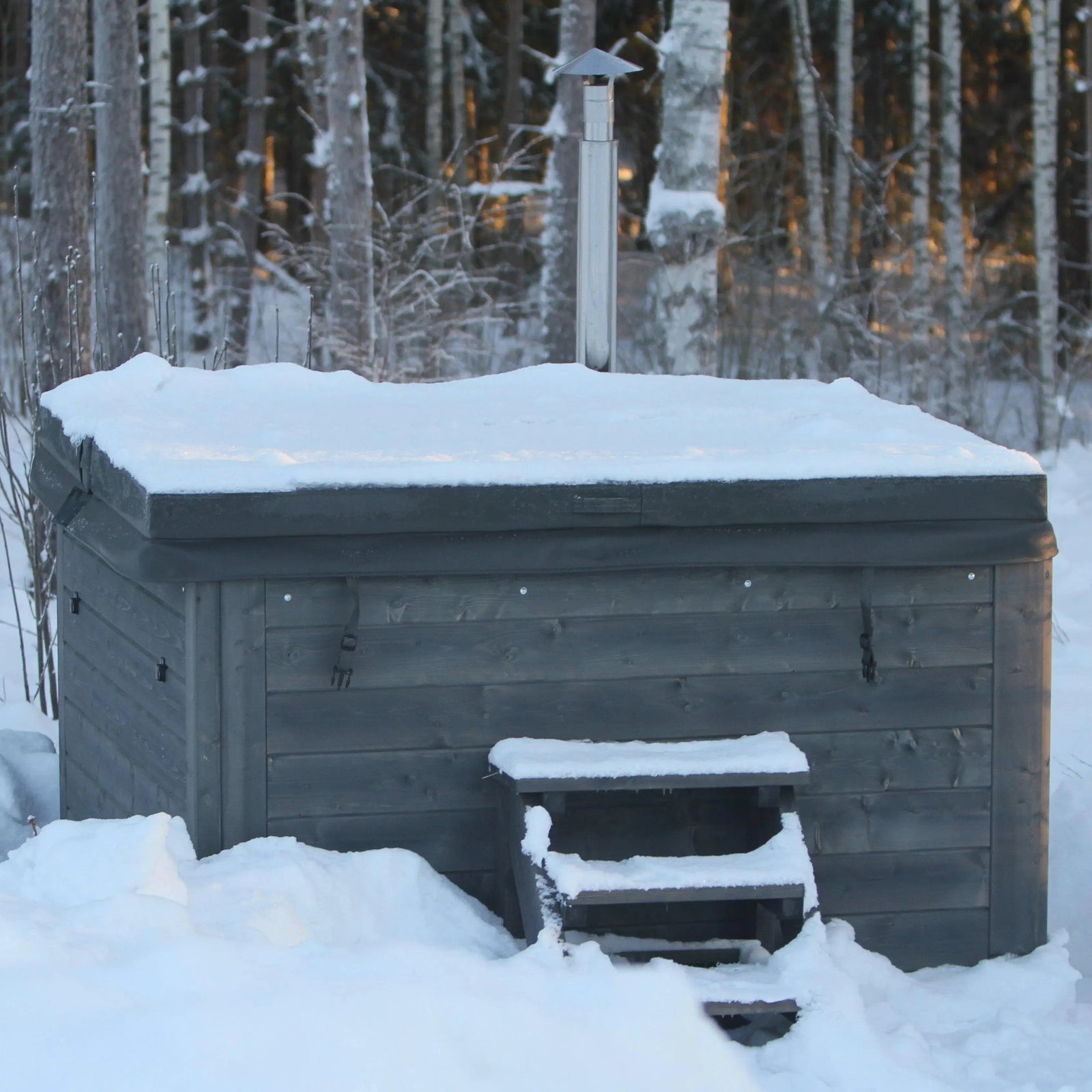 Nestled in a snowy forest, a wooden sauna with a snow-covered roof stands charmingly. A small set of wooden steps leads up to it, and a chimney protrudes from the top. Nearby, you can find the Rexener Polar Wooden Hot Tub - Made in Finland by Hot Tub Liverpool for an extra touch of relaxation amidst the serene surroundings filled with trees and snow.
