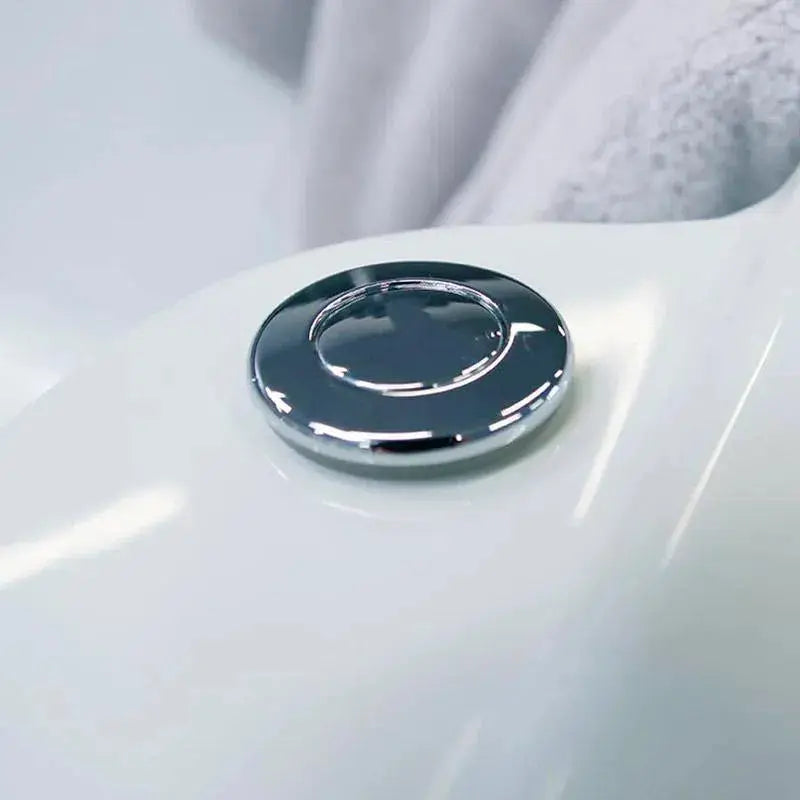 Close-up of a chrome-colored flush button on a white surface, possibly part of a toilet or sink. A soft, blurred grey towel is visible in the background. The image showcases the sleek and glossy design of the button, reflecting similar craftsmanship seen in Hot Tub Liverpool's Rexener Polar Wooden Hot Tub - Made in Finland.