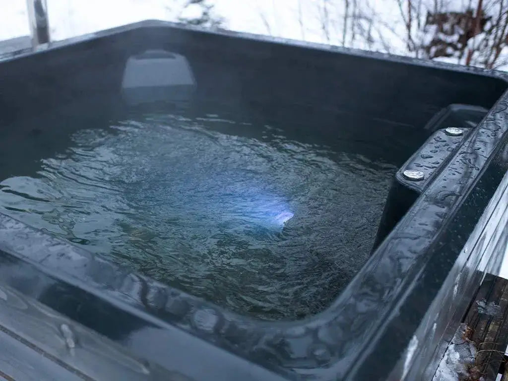 A handmade Rexener Polar Wooden Hot Tub - Made in Finland with clear, steaming water features subtle blue lighting. The wintery outdoor setting is evident with snow and bare branches in the background, adding a contrast between the warm water and cold surroundings. The Hot Tub Liverpool Rexener PR200 water heater ensures optimal warmth.