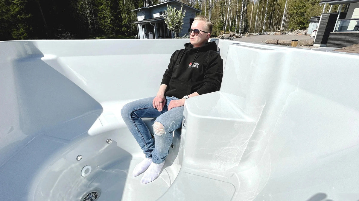 A person wearing sunglasses, a black hoodie, and ripped jeans is sitting in an empty Rexener Polar Wooden Hot Tub - Made in Finland by Hot Tub Liverpool outdoors. The person has their hands resting on their knees and is looking off into the distance, with trees and a house in the background.