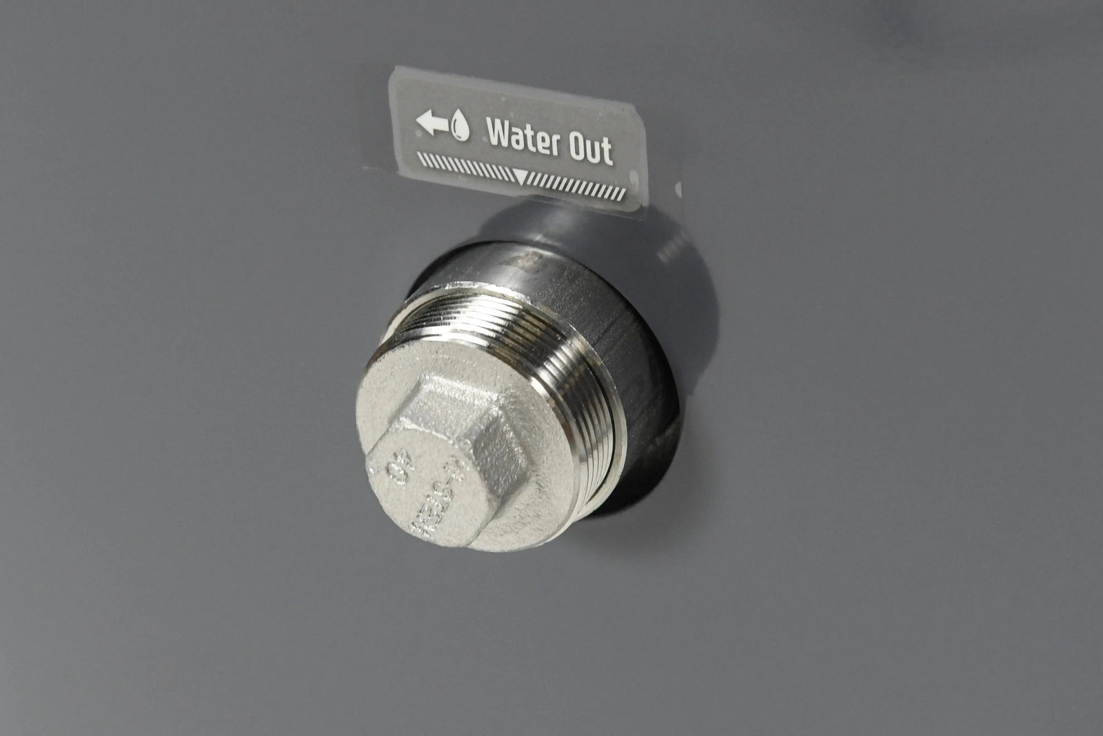 Close-up of a metallic water outlet plug on a gray surface, with a label above it reading "Water Out" along with an arrow pointing to the left. The plug is hexagonal and threaded, indicating where water exits, making it ideal for use with energy-efficient heaters like the Hot Tub Liverpool REXENER PR200 energy efficient hot tub water heater.