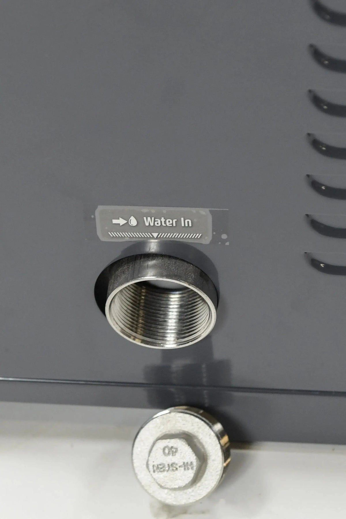 Close-up image of a metallic water inlet port on a machine, labeled "Water In" with an arrow pointing to the port—ideal for connecting Hot Tub Liverpool's REXENER PR200 energy efficient hot tub water heater. Below the port is a hexagonal metal bolt, all set against a grey painted surface.