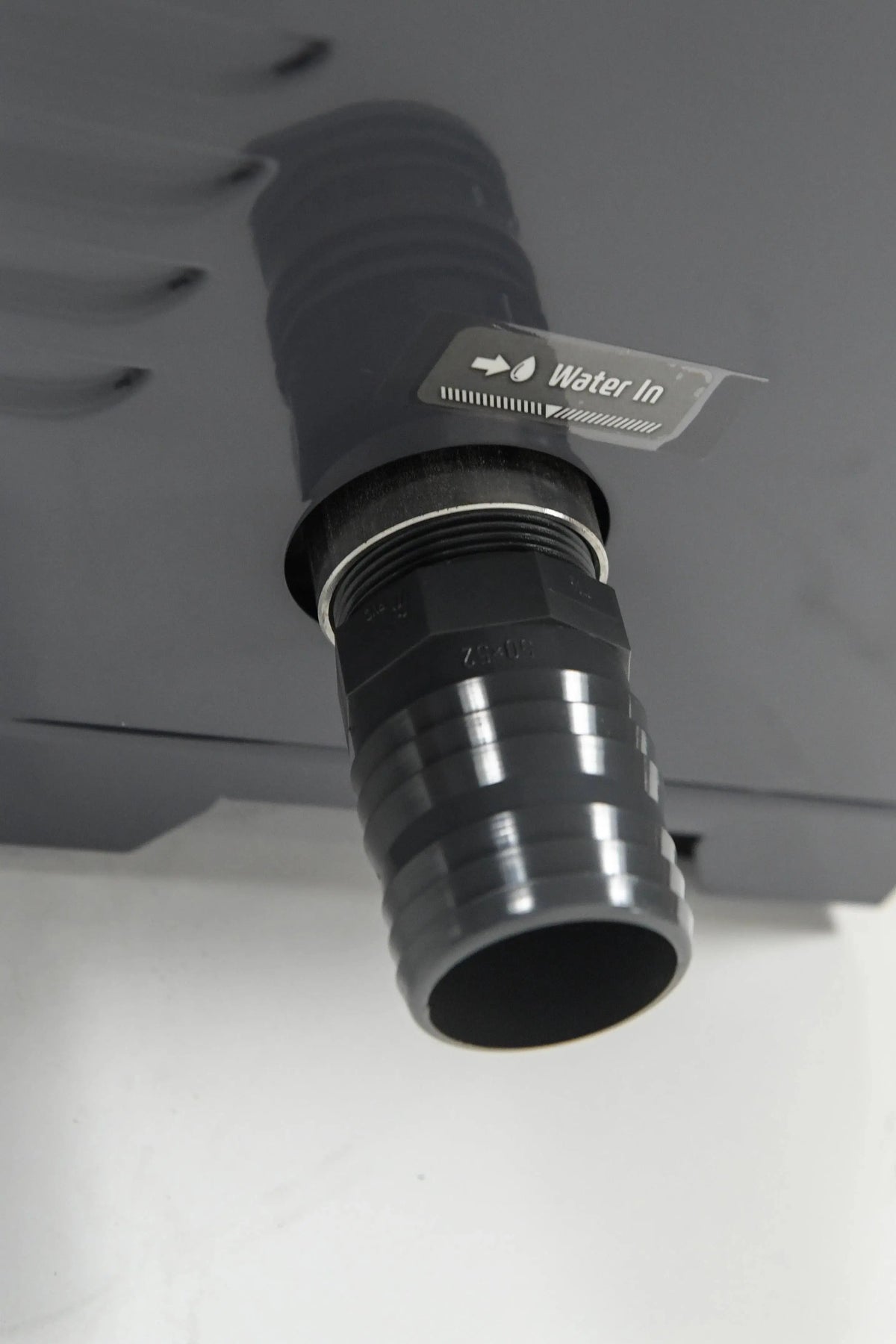 Close-up of a black water inlet pipe connected to a REXENER PR200 energy efficient hot tub water heater by Hot Tub Liverpool. A label above the pipe reads "Water In" with a direction arrow. The background is a light-colored surface.