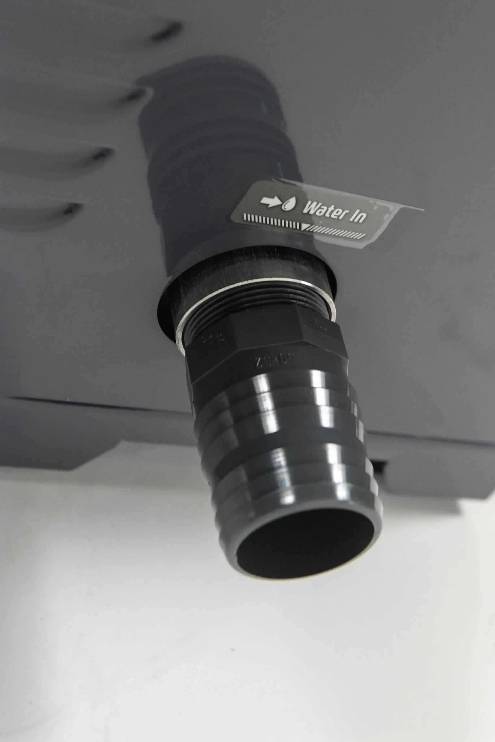 Close-up of a black water inlet pipe connected to a REXENER PR200 energy efficient hot tub water heater by Hot Tub Liverpool. A label above the pipe reads "Water In" with a direction arrow. The background is a light-colored surface.