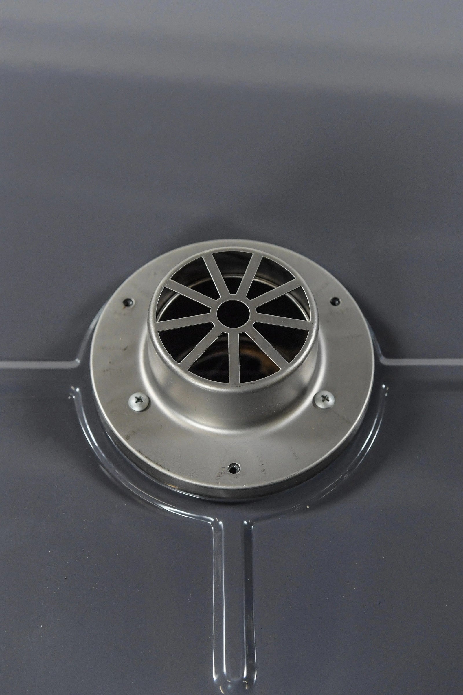 A close-up of a round metal vent embedded in a smooth, dark gray surface. The vent has a radial pattern with several evenly spaced spokes extending from the center hole. There are four screws securing the vent in place; it's reminiscent of the precision found in Hot Tub Liverpool's REXENER PR200 energy efficient hot tub water heater.