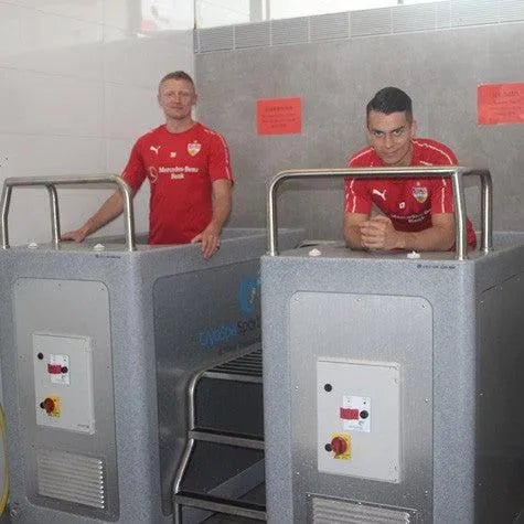 Two men are in CET CryoSpas Team CryoSpa Sport Ice Baths, possibly for athlete recovery, in a treatment room. They are both wearing red sports shirts with logos. One man is standing in a tub, and the other is leaning on the edge of another tub. The room has a modern, clinical appearance.