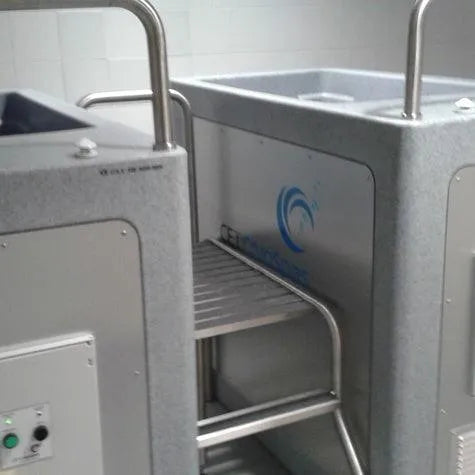 A Team CryoSpa Sport Ice Bath designed for cryotherapy sessions stands in a clean, tiled space. The grey cabin features a blue logo and safety rails with a step stool for easy entry. Control panels are visible on the side, indicating temperature and settings adjustments, ideal for athlete recovery.