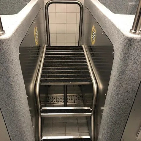 This image shows a public foot washing station designed for athlete recovery. It features a silver metal grate for foot support, with steps leading up to it. The background has a tiled wall and the station is framed by gray and metal surfaces. An overhead metal rail provides stability, reminiscent of CET CryoSpas' Team CryoSpa Sport Ice Bath facilities.